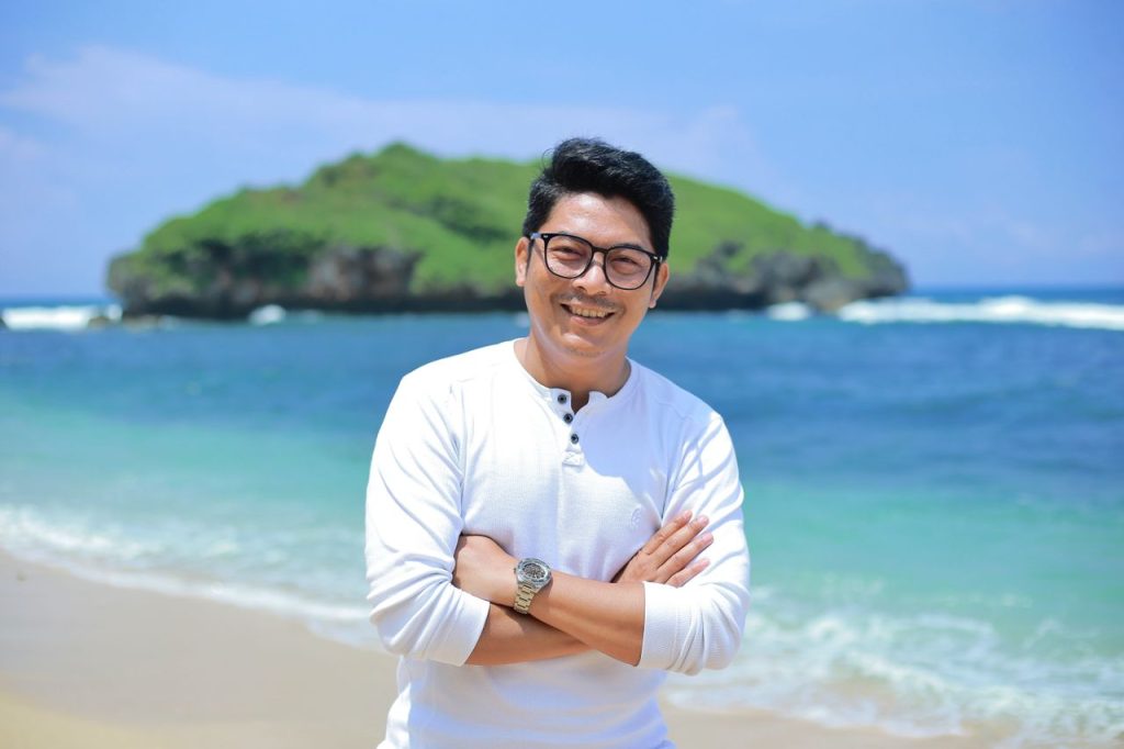 Cheerful man with glasses standing on a sunny beach with a scenic island backdrop.